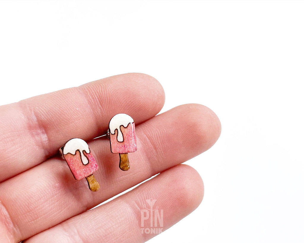 A pair of small stud earrings shaped like popsicles, held between fingers against a plain white background. The earrings have pink bodies, white dripping tops, and brown wooden sticks.