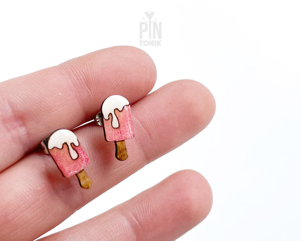 A pair of small stud earrings shaped like popsicles, shown resting on a person's fingers against a plain white background. The earrings have pink bodies, white dripping tops, and brown wooden-style sticks.