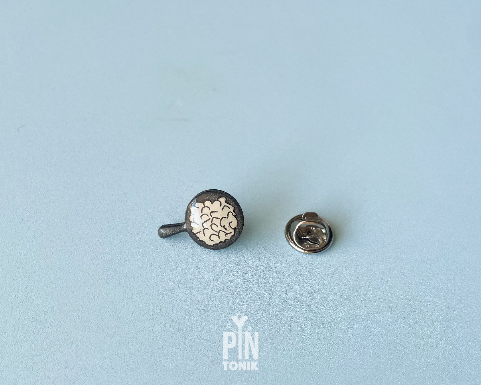 Hand-painted wooden pin shaped like a black frying pan containing a sugar-dusted kaiserschmarrn pancake, shown next to a stainless steel clutch back on a light blue background.