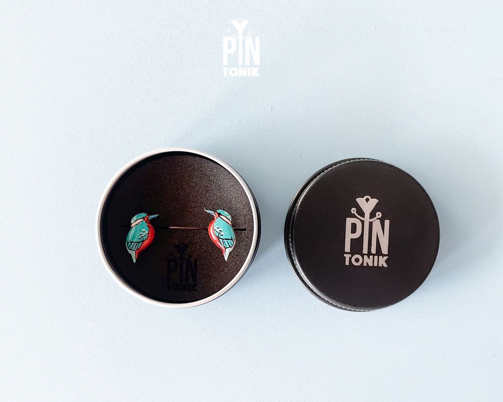 A pair of colorful bird stud earrings displayed inside an open round metal tin next to its lid, both featuring the PIN TONIK brand logo, on a light blue background.