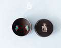 A pair of colorful bird stud earrings displayed inside an open round metal tin next to its lid, both featuring the PIN TONIK brand logo, on a light blue background.