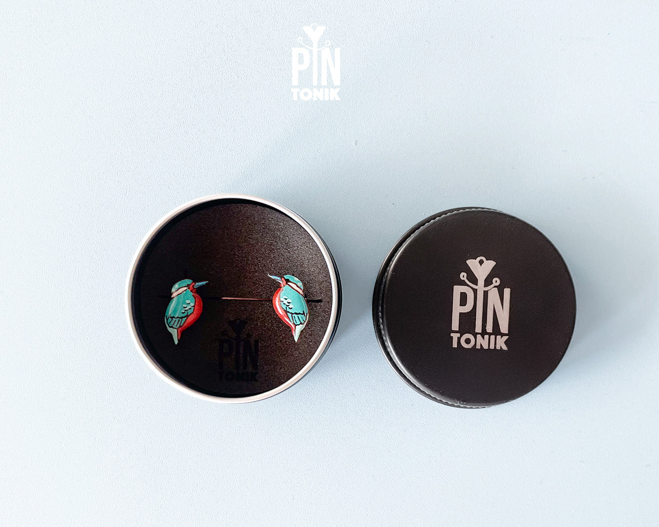 A pair of colorful bird stud earrings displayed inside an open round metal tin next to its lid, both featuring the PIN TONIK brand logo, on a light blue background.