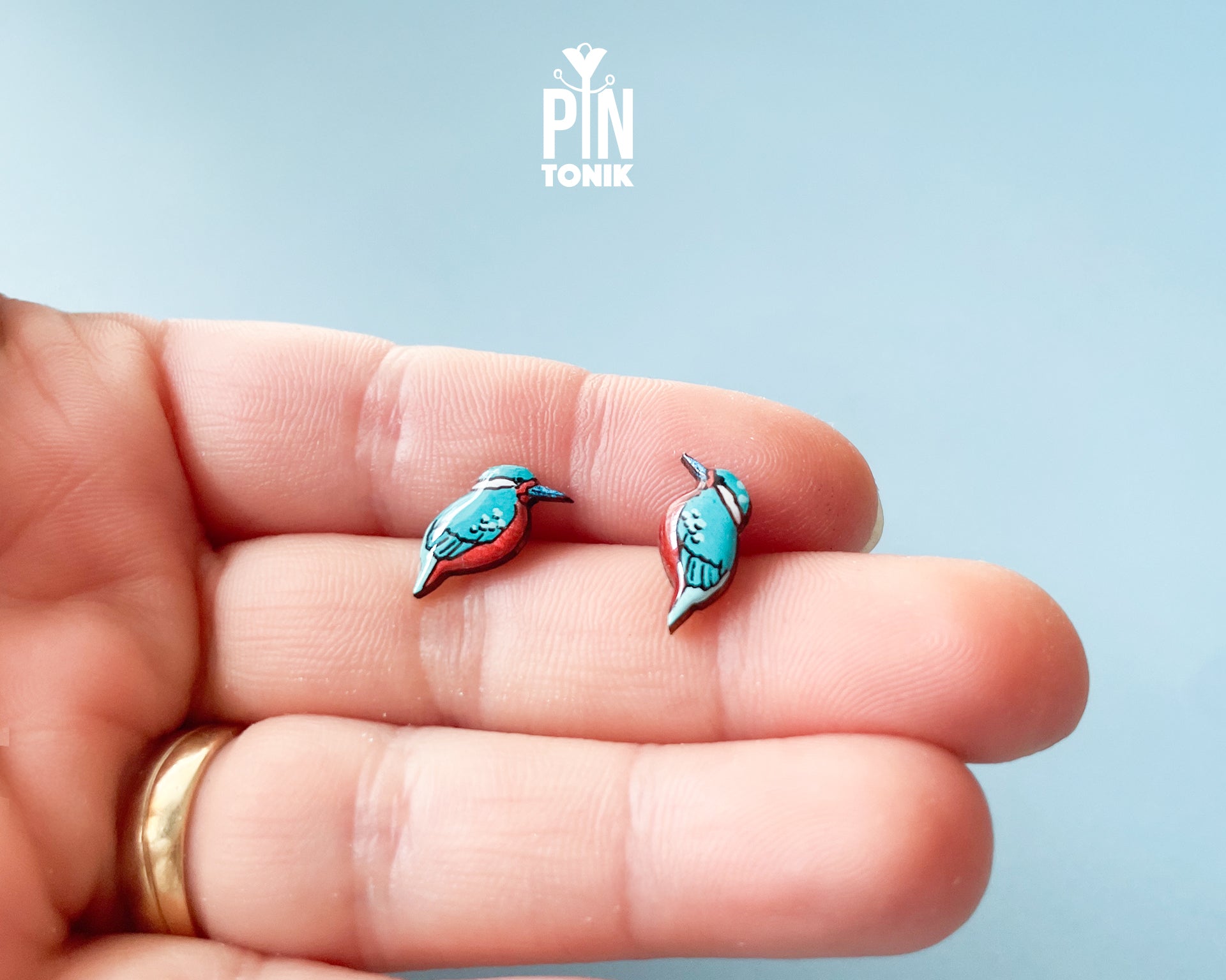 Two small bird-shaped pins in turquoise blue and pink are displayed on an open hand, with a gold ring visible on the wrist against a soft light blue background. The brand logo 'PIN TONIK' is shown at the top center.