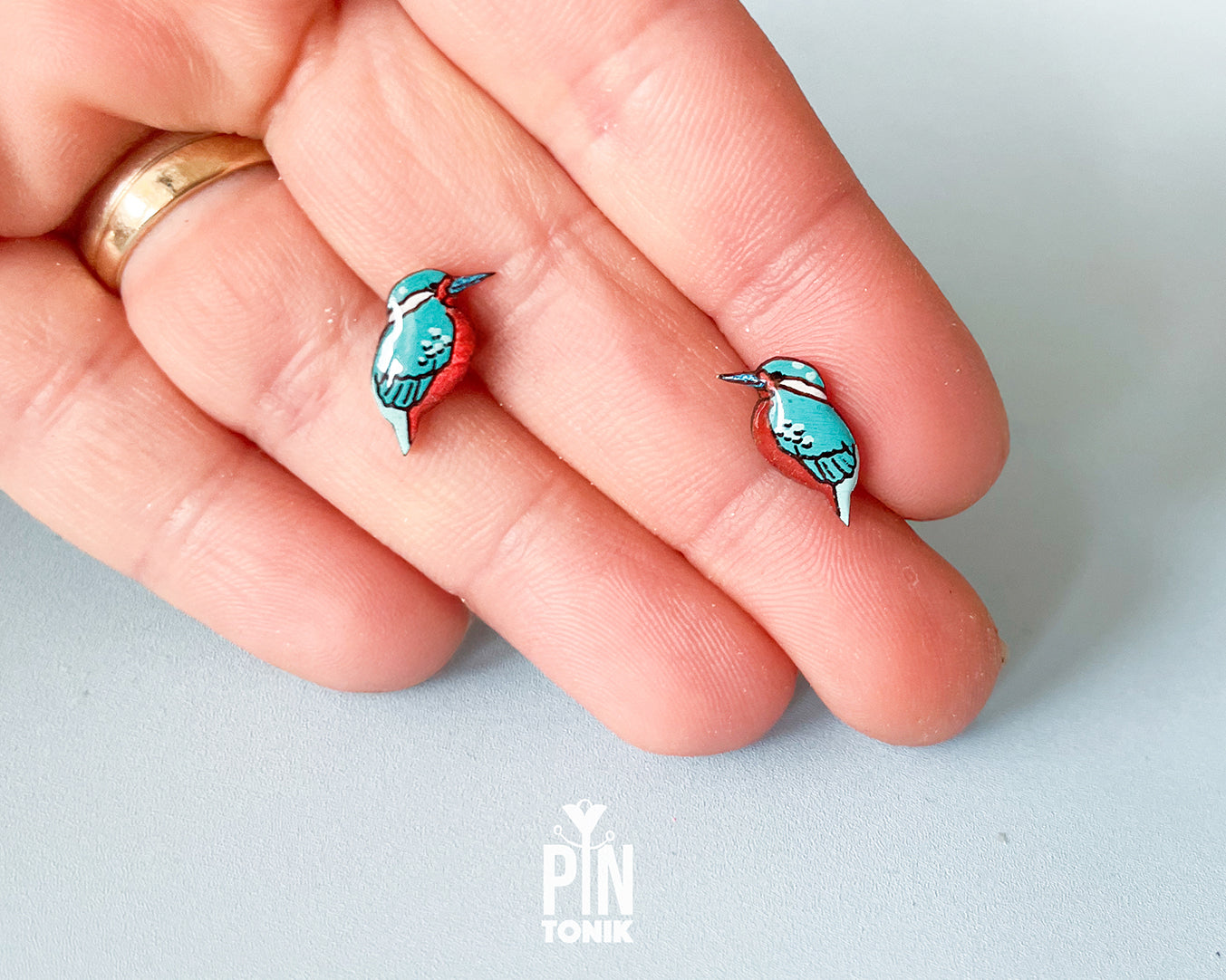 A close-up of a hand holding two small bird-shaped earrings with turquoise blue, red, and white enamel details, with a gold ring on the finger for scale. The brand name 'PIN TONIK' is visible as a watermark at the bottom.
