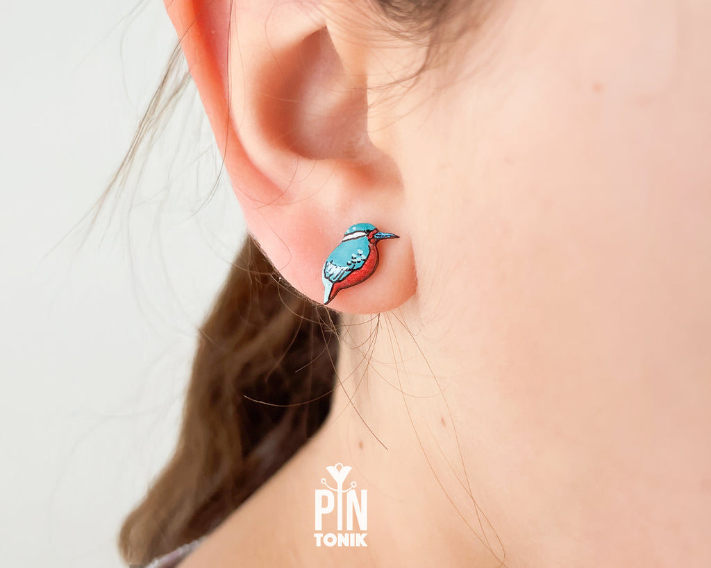 A close-up of a person's ear wearing a small bird-shaped stud earring in turquoise blue, red, and white, with the brand name 'PIN TOMIK' visible as a watermark at the bottom.
