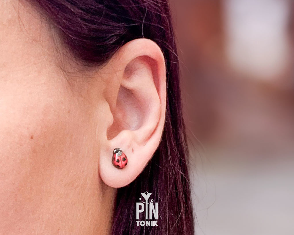 A close-up of a person's ear wearing a small red ladybug stud earring with black spots and tiny silver accents. The background is a soft, blurred neutral tone, and a 'PIN TONIK' watermark is visible at the bottom.