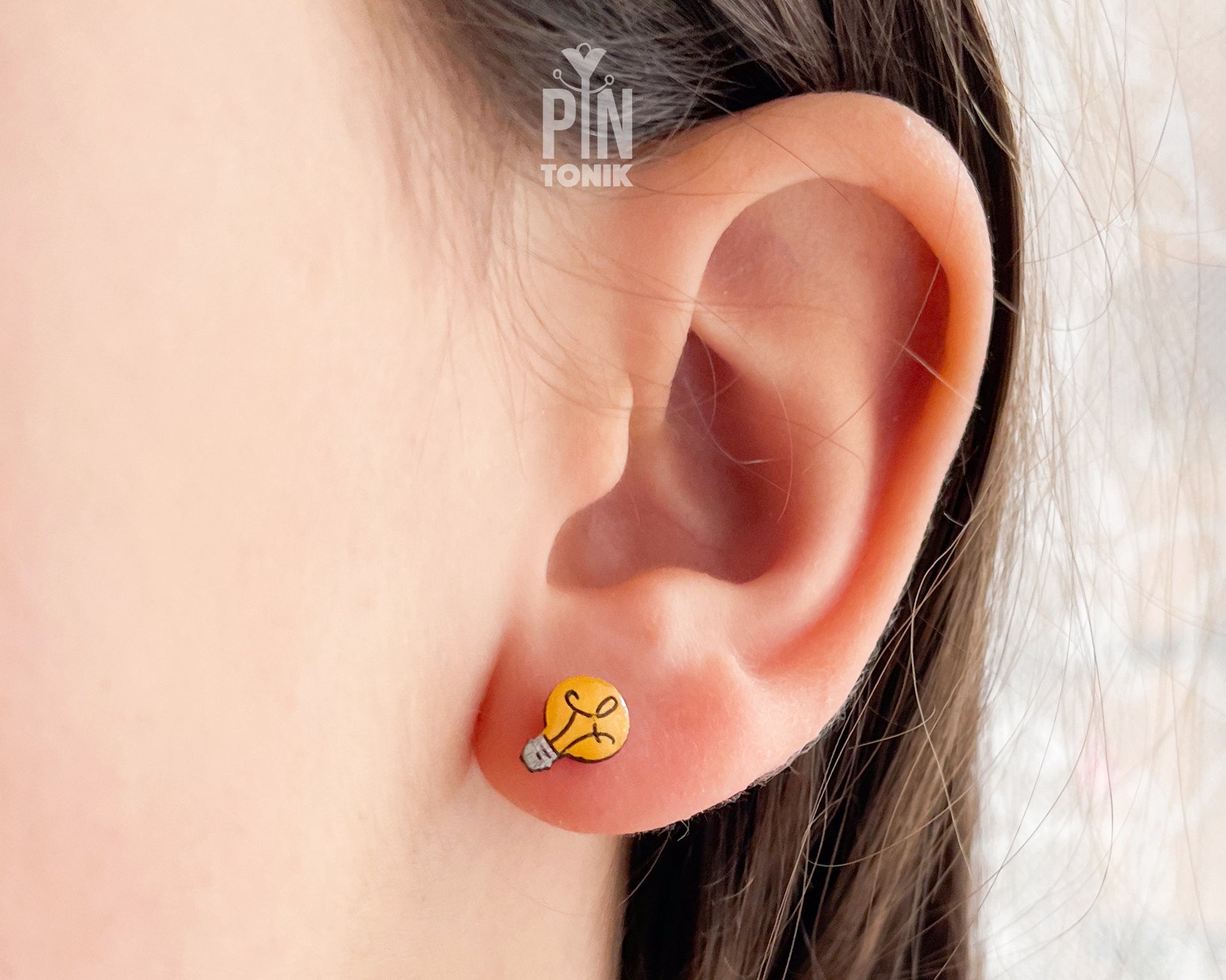 A close-up of a person's ear wearing a small yellow stud earring shaped like a lightbulb with a black lines and a silver-gray tip. A white 'PIN TONIK' watermark is visible near the top.