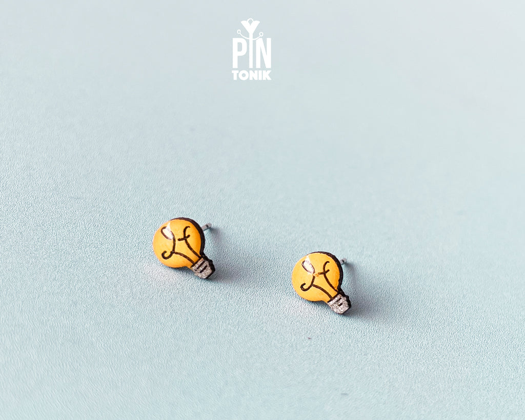 A pair of lightbulb-shaped wooden stud earrings in yellow with black line details and stainless steel posts, displayed on a light blue-gray textured surface. The brand logo 'PIN TONIK' is visible at the top center.