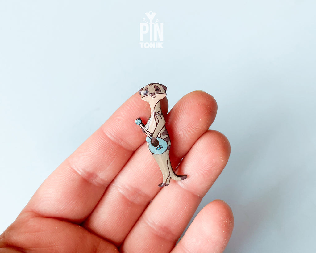 Front View: A detailed wooden lapel pin of a whimsical meerkat playing a blue guitar, shown next to its gold butterfly clutch backing on a minimal light blue background.
Angled View: A high-quality wooden art pin featuring a musical meerkat illustration, displayed with its gold butterfly backing on a dual-tone yellow and blue background.