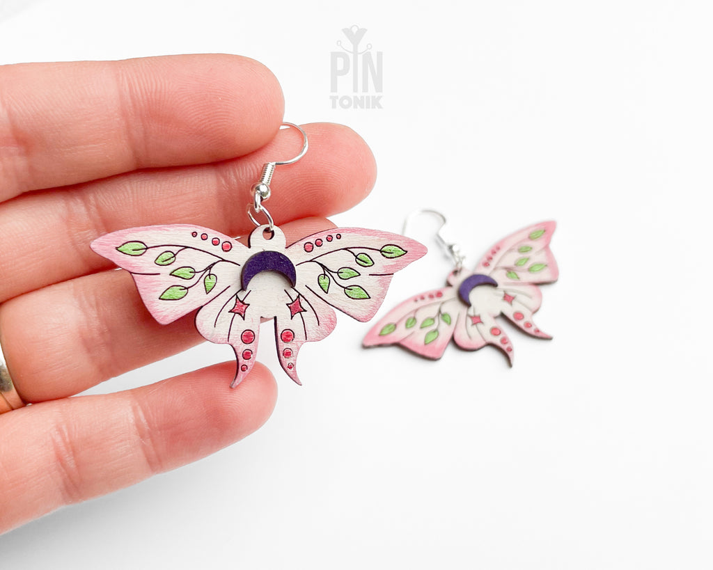 Hand-painted wooden Luna Moth dangle earrings featuring pink wings, green leaf patterns, and purple moon phases on sterling silver hooks
