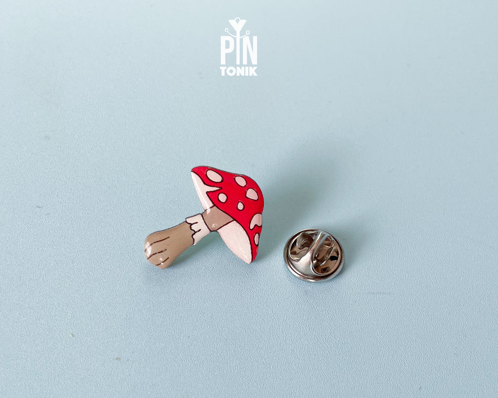 Hand-painted wooden mushroom pin with a bright red cap and cream spots, shown next to a stainless steel clutch back on a light blue background.
