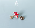 Hand-painted wooden mushroom pin with a bright red cap and cream spots, shown next to a stainless steel clutch back on a light blue background.