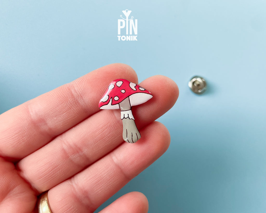 Hand-painted wooden mushroom pin with a bright red cap and cream spots, shown next to a stainless steel clutch back on a light blue background.