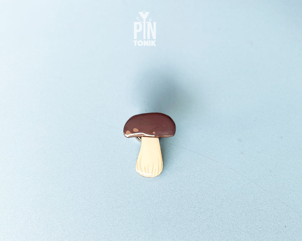 Hand-painted wooden porcini mushroom pin with a dark brown cap and cream-colored stem, shown on a light blue background