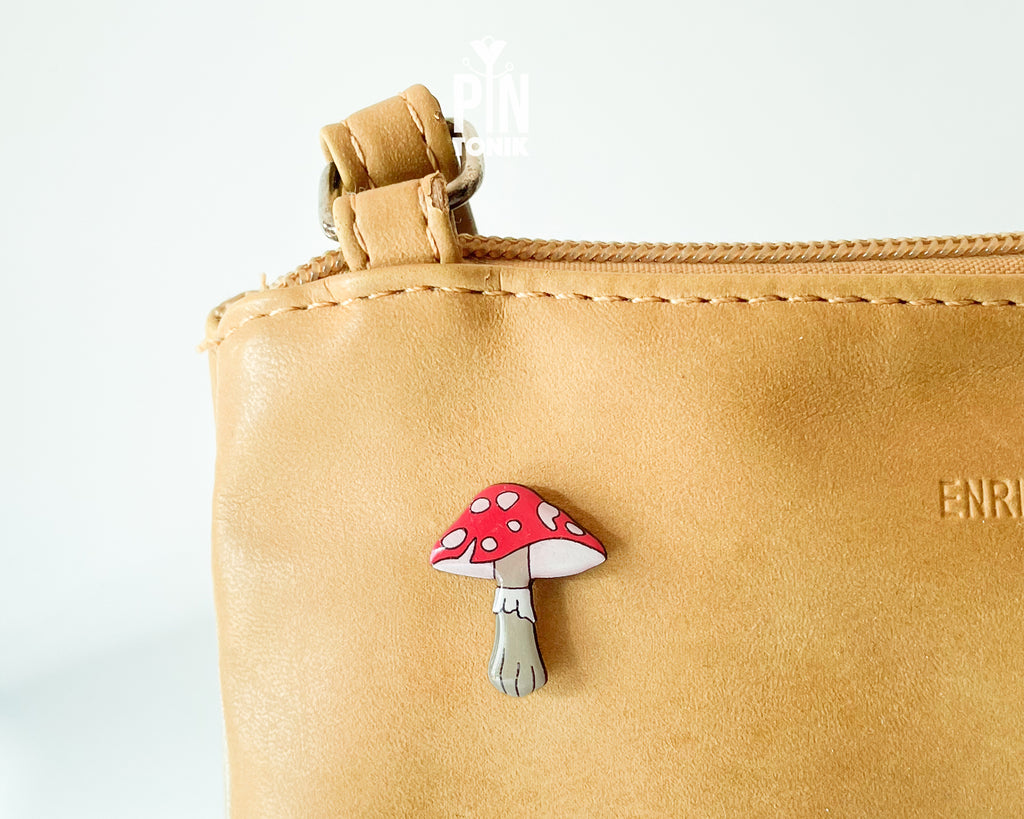 Hand-painted wooden mushroom pin with a bright red cap and cream spots, shown next to a stainless steel clutch back on a light blue background.