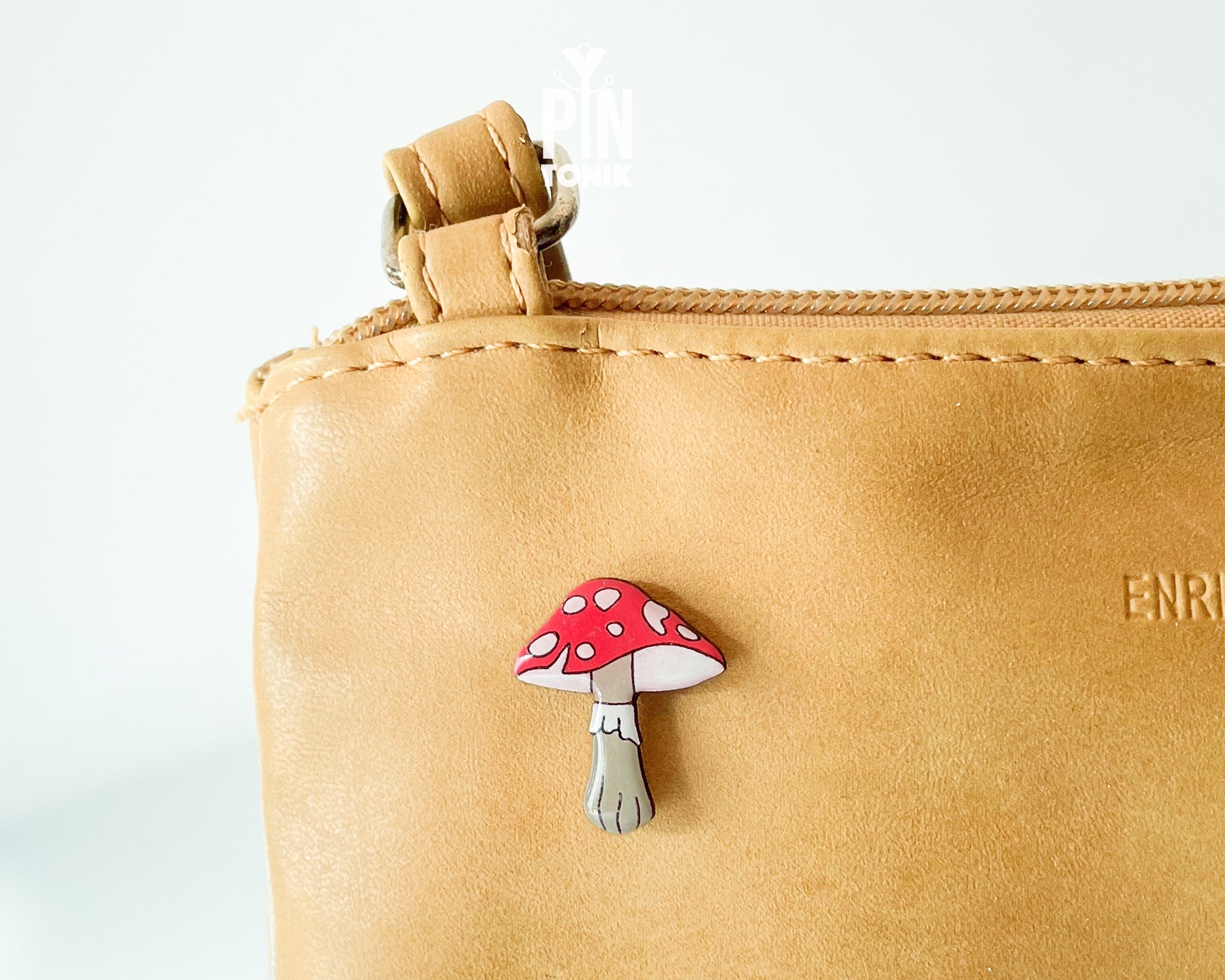 Hand-painted wooden mushroom pin with a bright red cap and cream spots, shown next to a stainless steel clutch back on a light blue background.