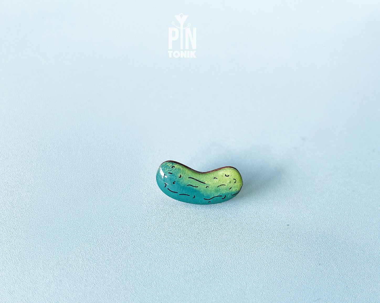 Hand-painted wooden pickle pin with light and dark green gradients and detailed textures, shown on a light blue background.