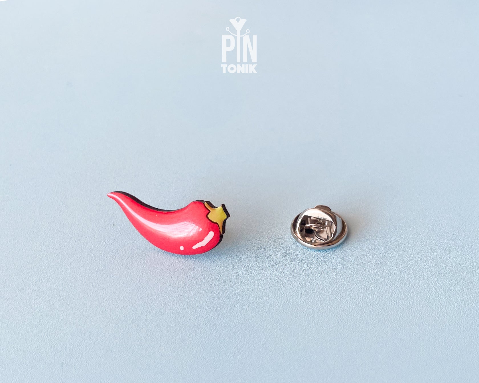 Hand-painted wooden red chili pepper pin with a glossy finish and a yellow stem, shown next to its stainless steel clutch back on a light blue-grey background