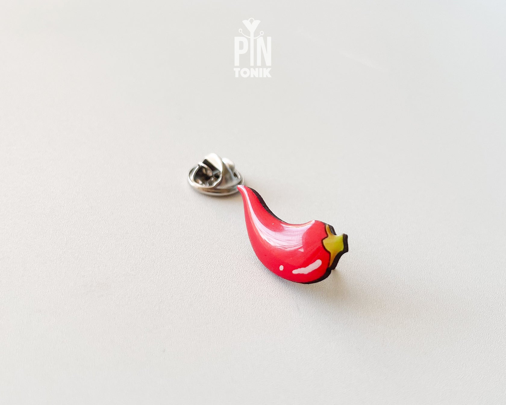 Hand-painted wooden red chili pepper pin with a glossy finish and a yellow stem, shown next to its stainless steel clutch back on a light blue-grey background