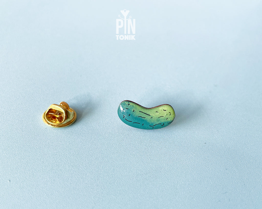 Hand-painted wooden pickle pin with light and dark green gradients and detailed textures, shown on a light blue background.