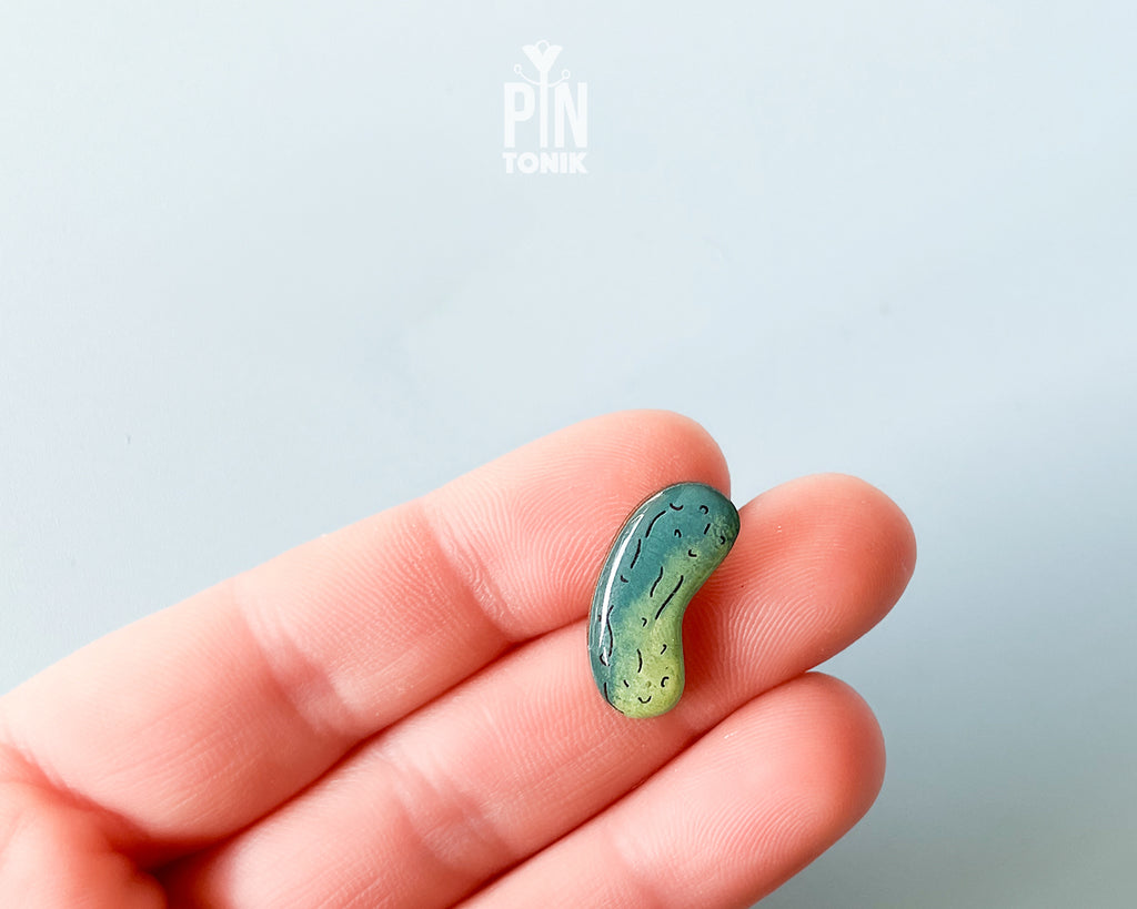 Hand-painted wooden pickle pin with light and dark green gradients and detailed textures, shown on a light blue background.