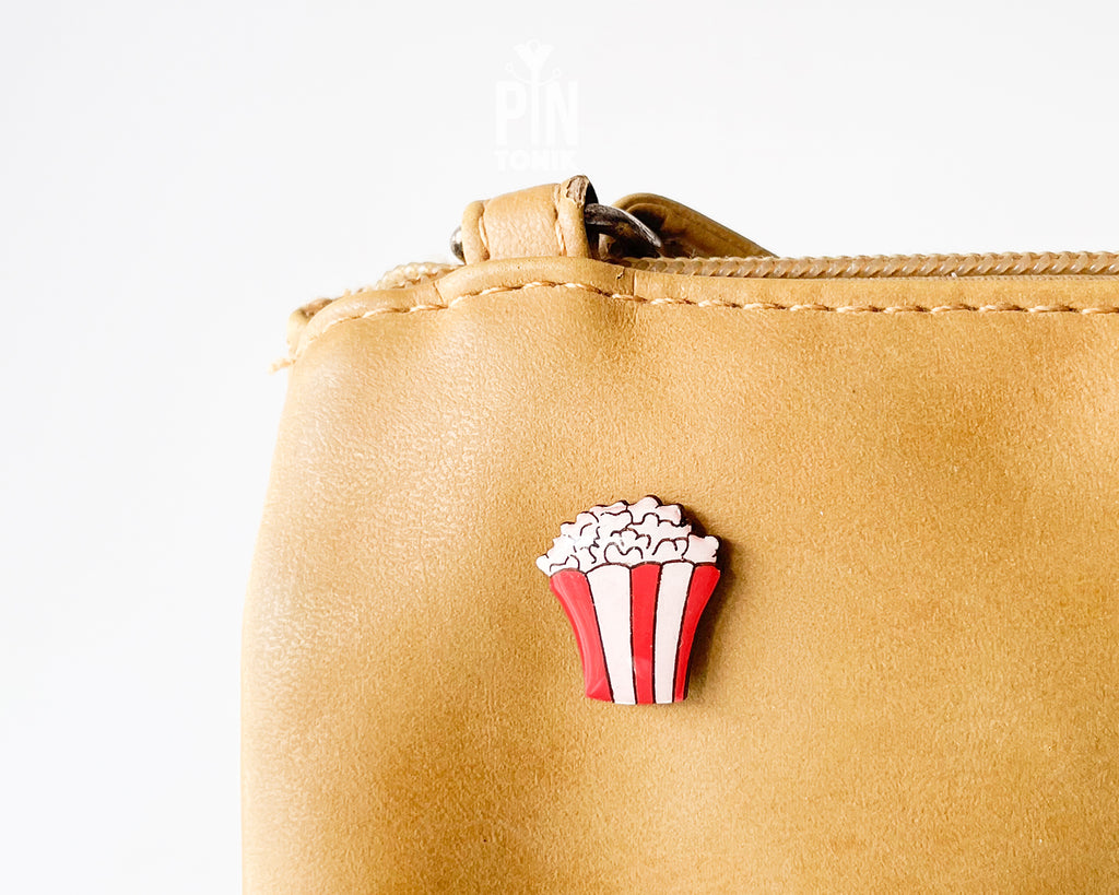 Hand-painted wooden popcorn bucket pin with red and white stripes and glossy white popcorn kernels, shown next to a stainless steel clutch back on a light blue background.