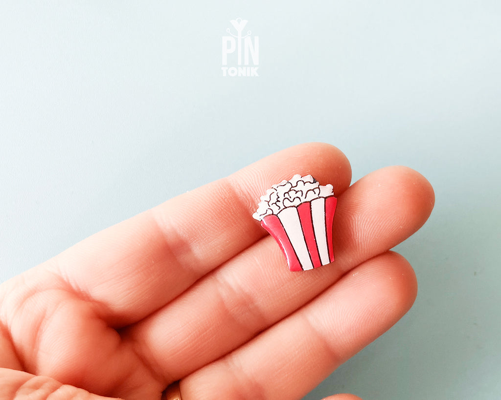 Hand-painted wooden popcorn bucket pin with red and white stripes and glossy white popcorn kernels, shown next to a stainless steel clutch back on a light blue background.