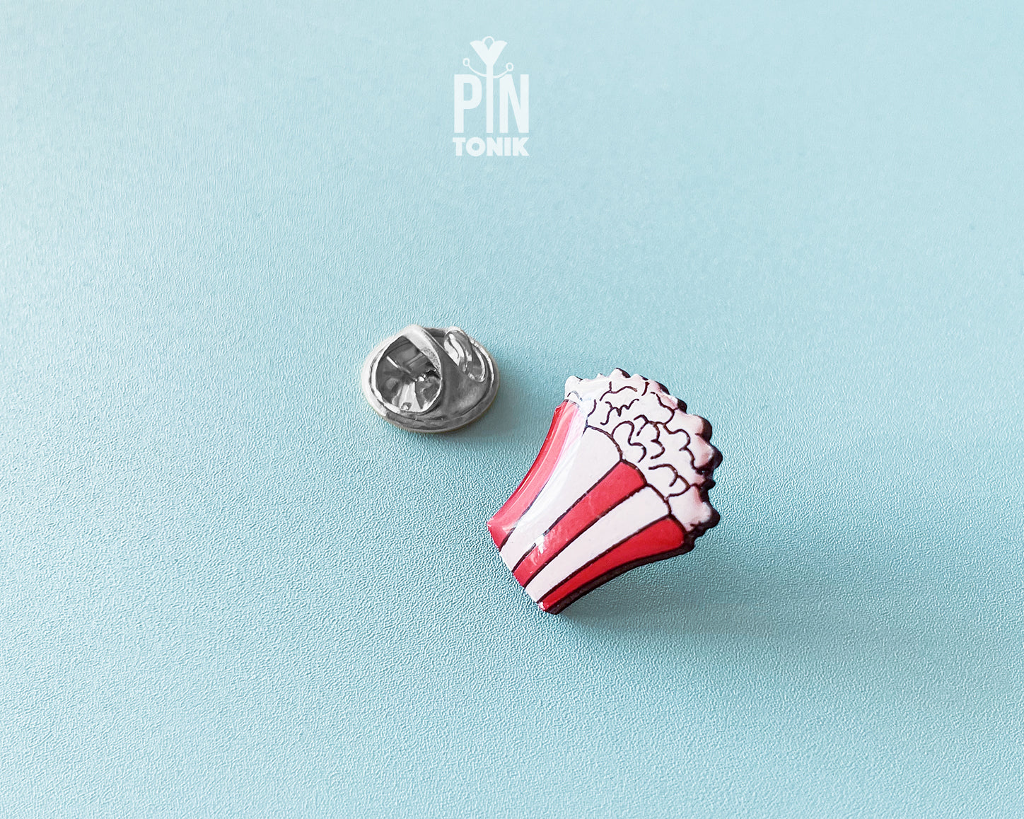 Hand-painted wooden popcorn bucket pin with red and white stripes and glossy white popcorn kernels, shown next to a stainless steel clutch back on a light blue background.
