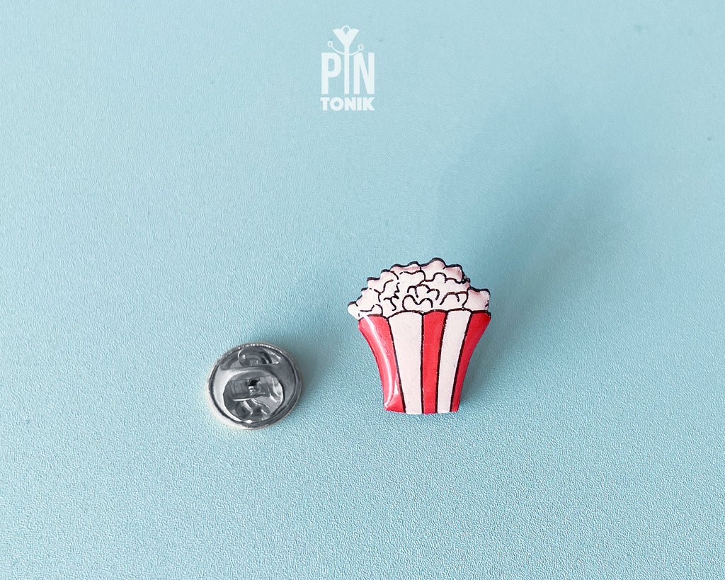 Hand-painted wooden popcorn bucket pin with red and white stripes and glossy white popcorn kernels, shown next to a stainless steel clutch back on a light blue background.