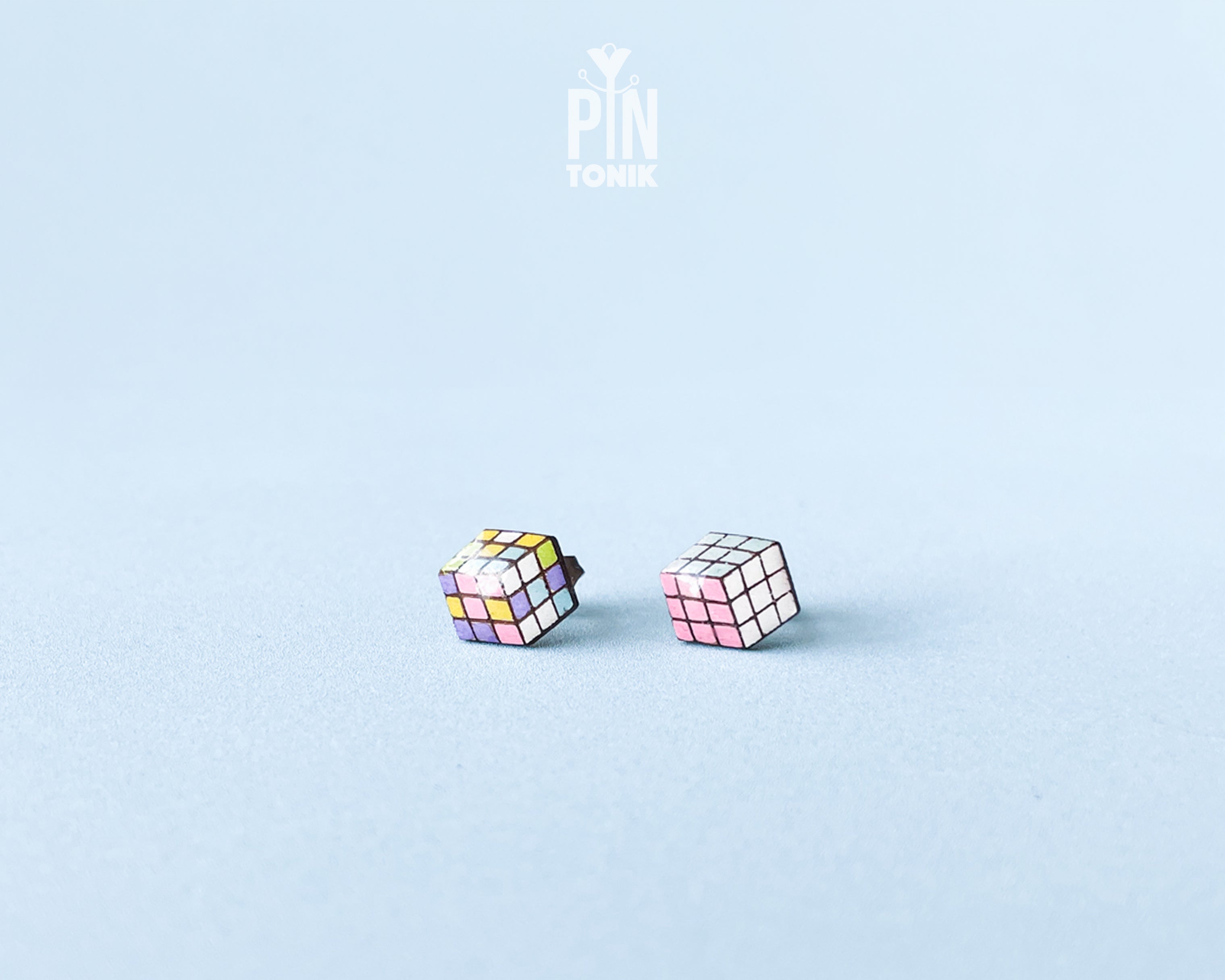 A pair of cube-shaped wooden stud earrings with colorful puzzle-cube faces, displayed on a light blue background with a 'PIN TONIK' logo at the top.