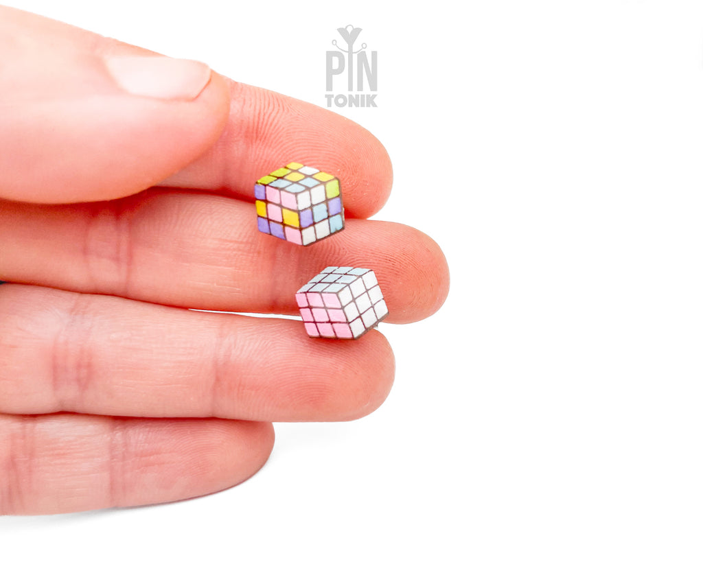 Two miniature cube-shaped earrings with a multicolor puzzle design are held between a person's fingers against a plain white background.