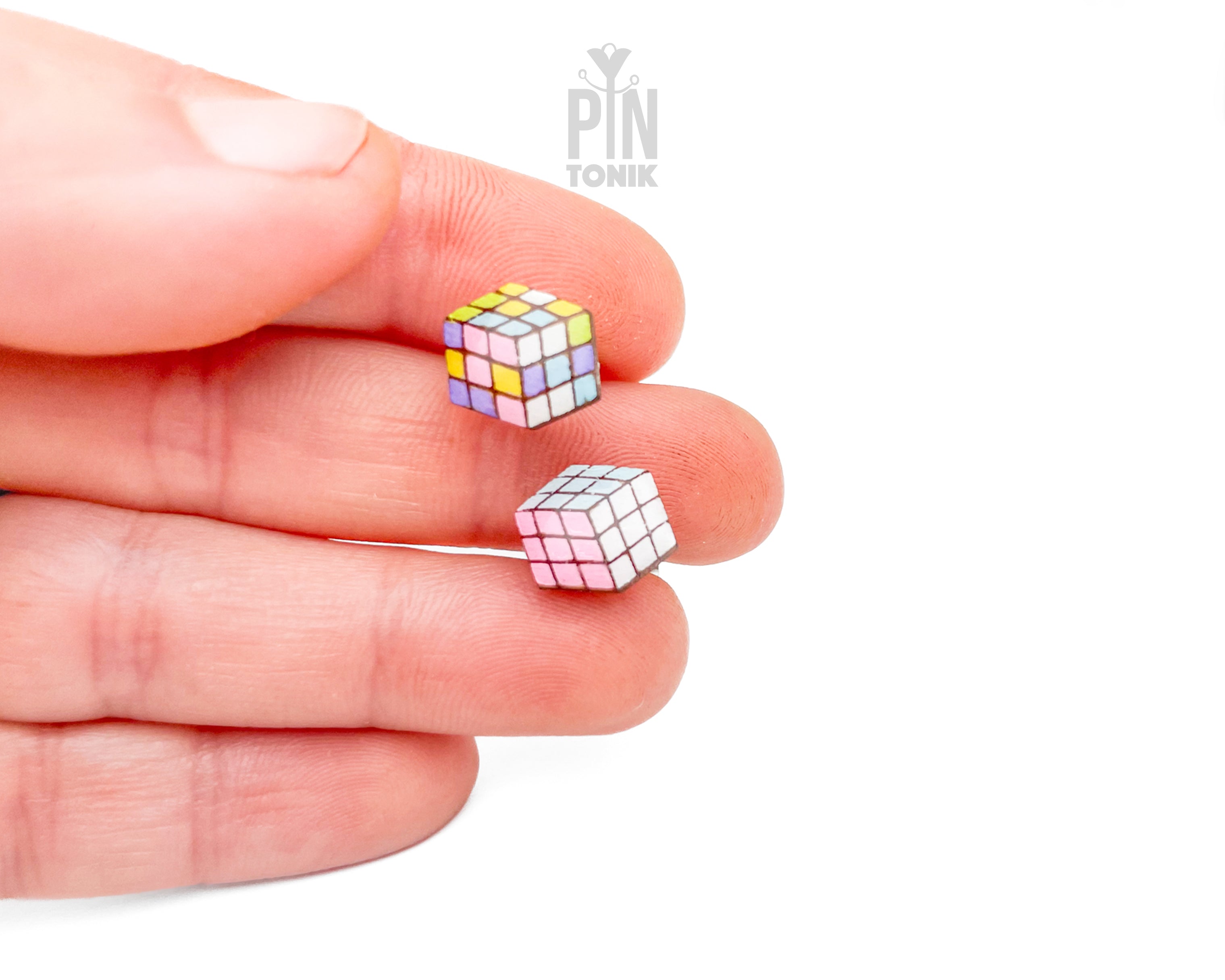Two miniature cube-shaped earrings with a multicolor puzzle design are held between a person's fingers against a plain white background.
