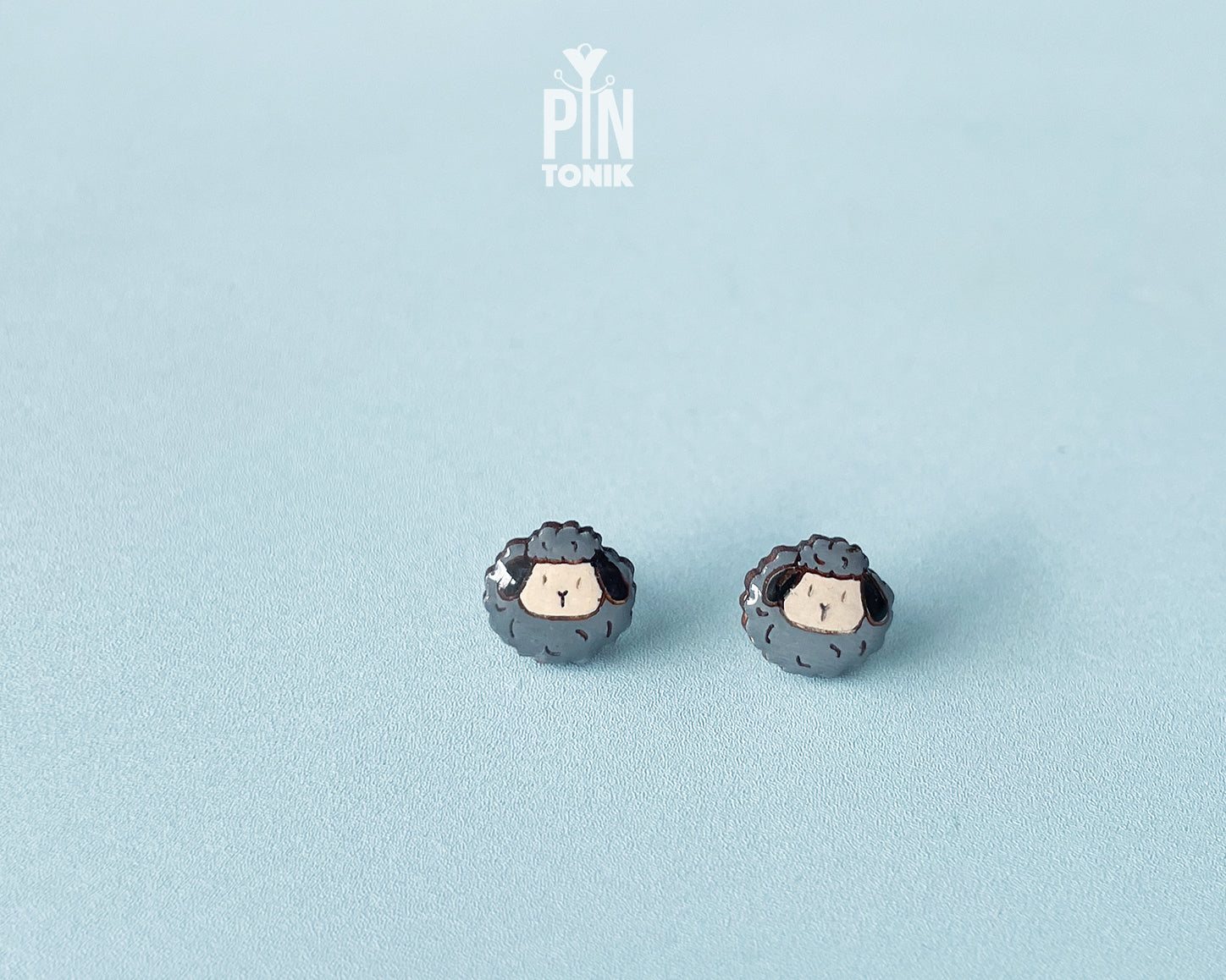 A pair of small stud earrings shaped like cute sheep with dark gray wool and cream-colored faces, displayed on a light blue textured background.