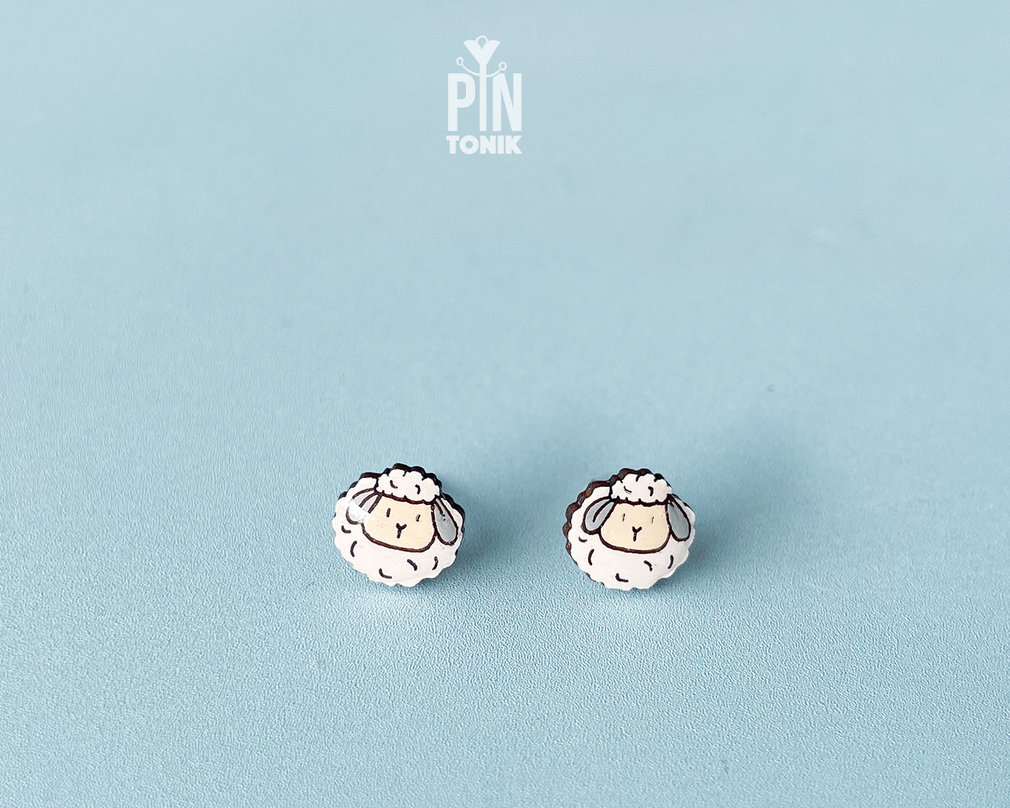 A pair of small stud earrings shaped like cartoon sheep, shown on a light blue background. The sheep have white woolly bodies with black outlines and beige faces.
