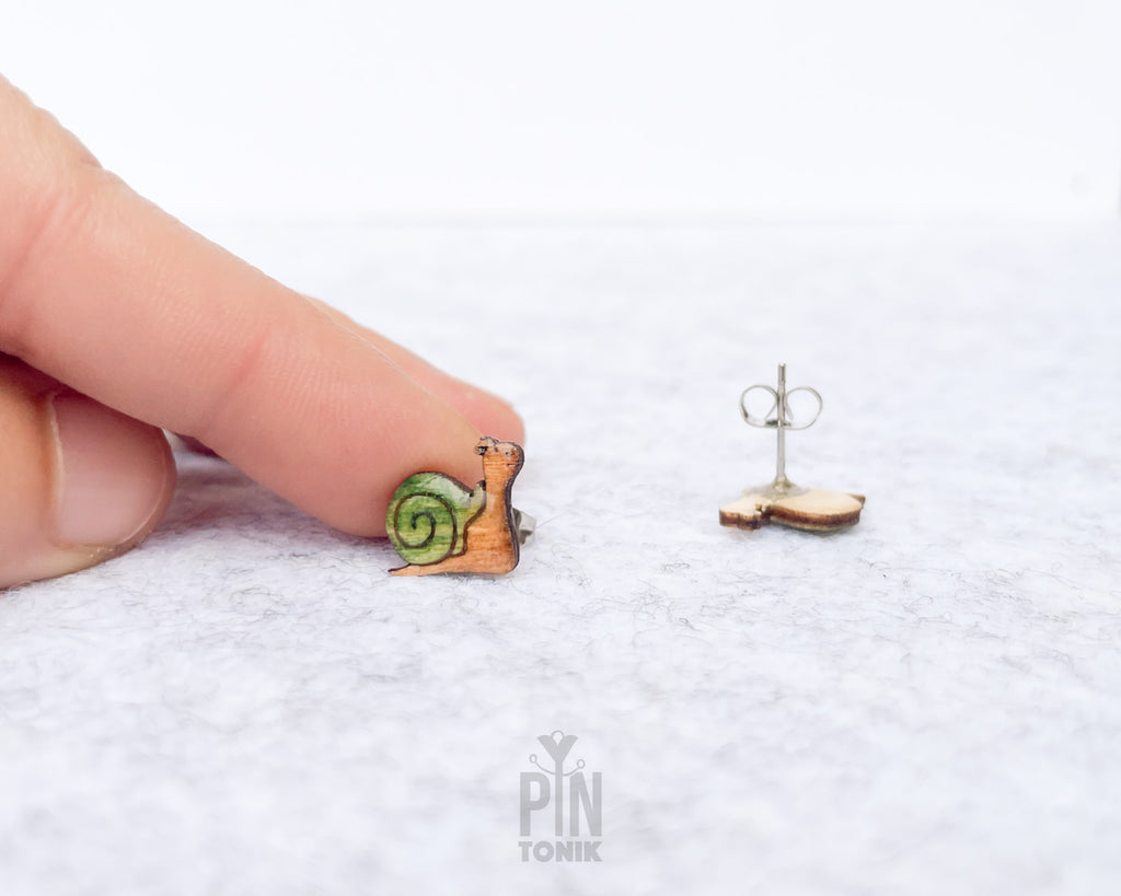 A close-up of a tiny gold snail stud earring with a green and yellow shell, shown next to a human finger for scale, with the back of the earring on a white textured surface.