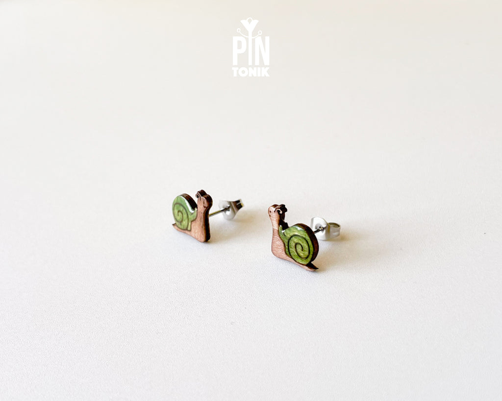 A pair of small stud earrings shaped like snails, with green spiral shells and copper-toned bodies, photographed on a plain light background. A small 'PIN TOMIK' mark appears near the top center.