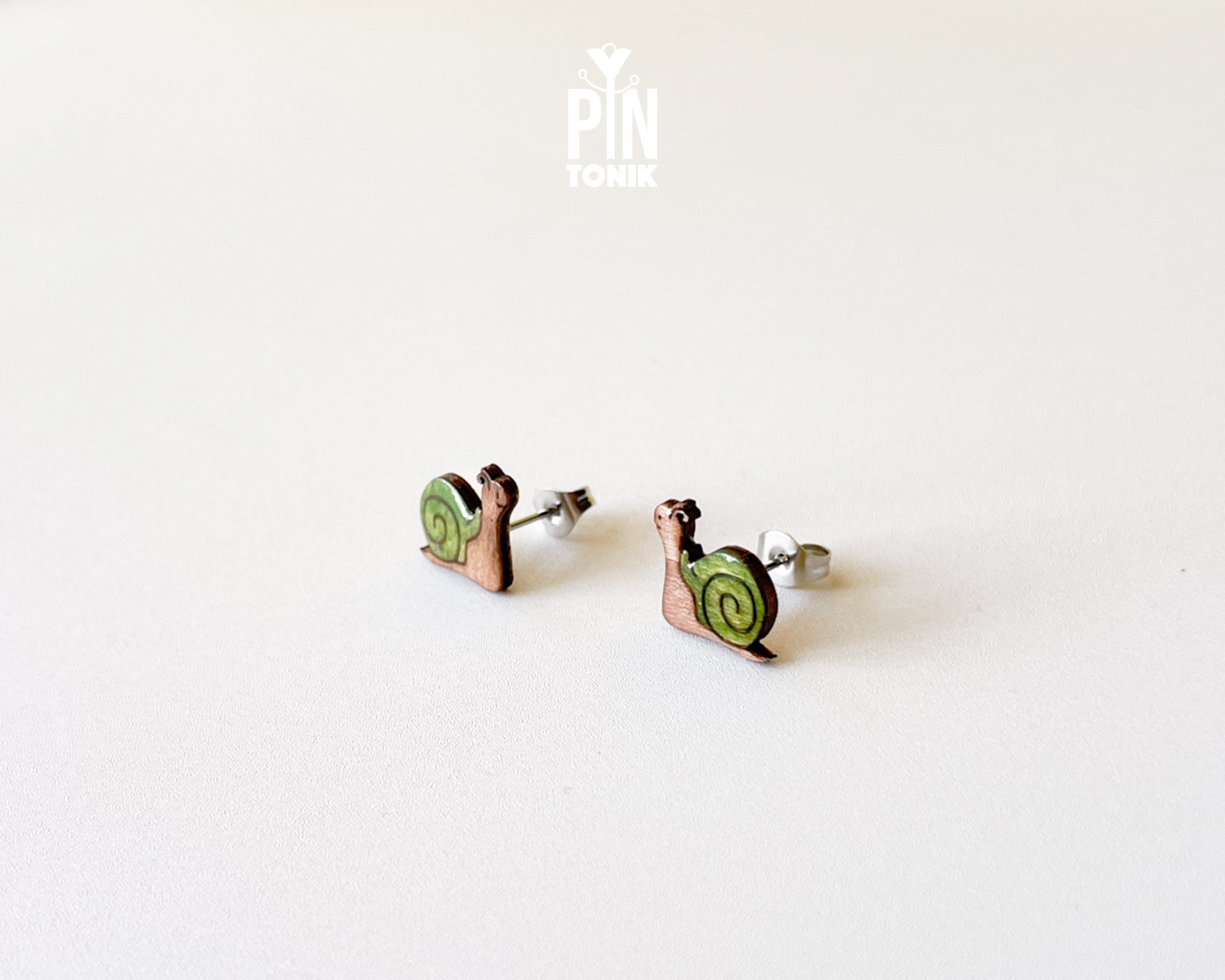 A pair of small stud earrings shaped like snails, with green spiral shells and copper-toned bodies, photographed on a plain light background. A small 'PIN TOMIK' mark appears near the top center.