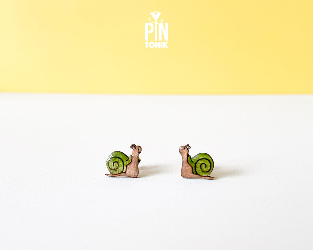 A pair of small snail stud earrings with green shells and pinkish-brown bodies, displayed on a white surface against a yellow background with a 'PIN TOMIK' logo at the top.