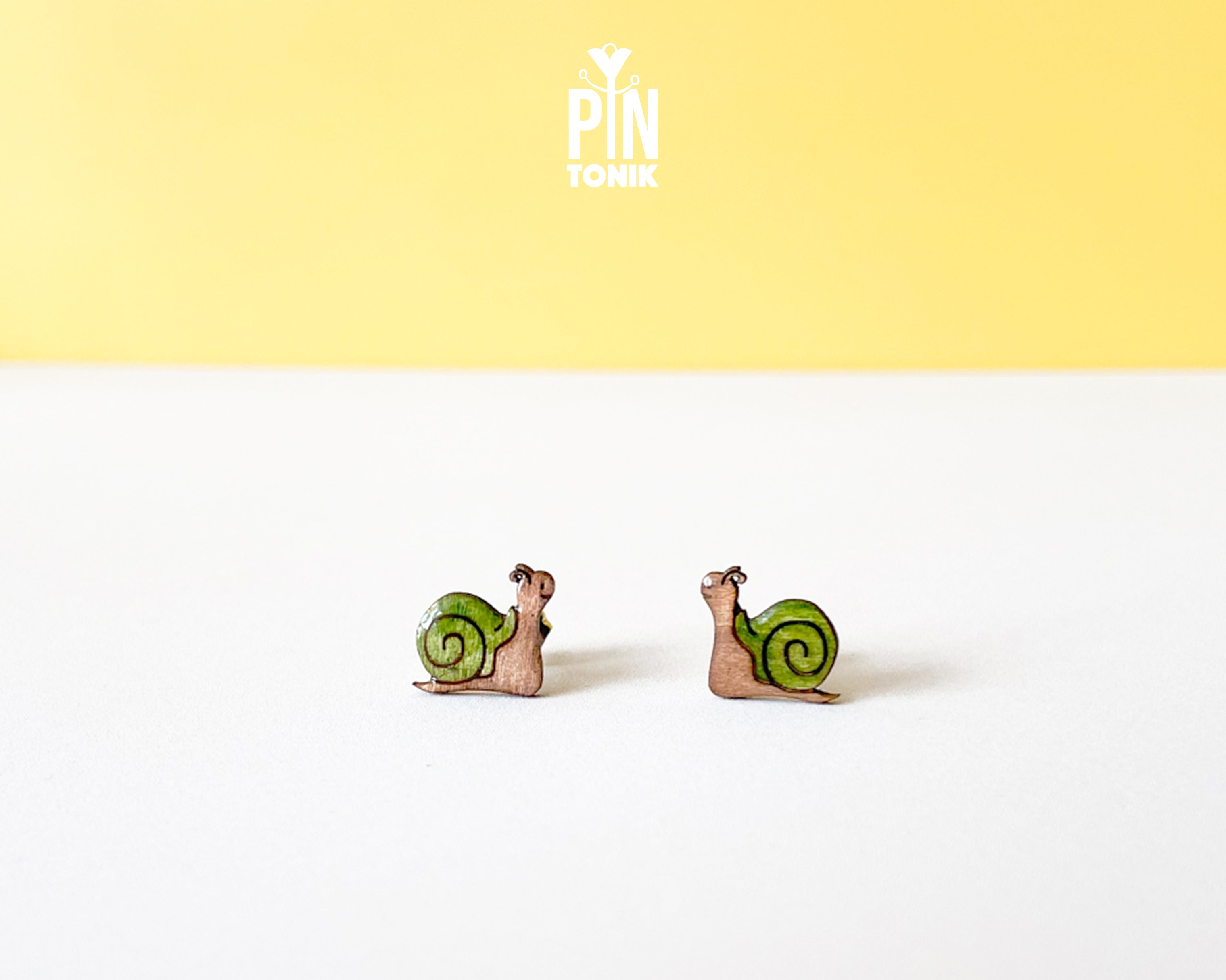 A pair of small snail stud earrings with green shells and pinkish-brown bodies, displayed on a white surface against a yellow background with a 'PIN TOMIK' logo at the top.