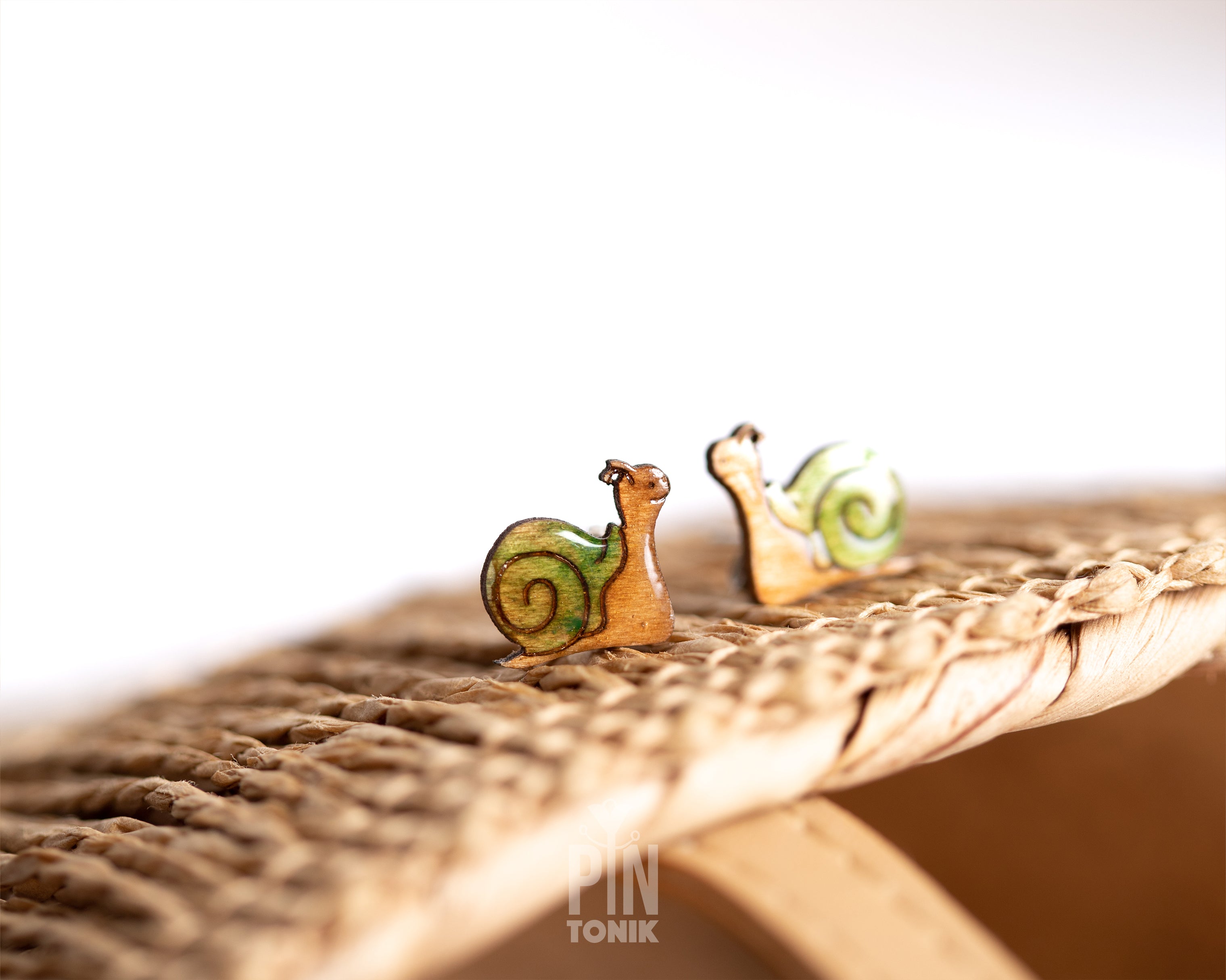 Two small snail-shaped earrings with green and yellow shells and tan bodies, resting on a woven straw surface against a bright white background.