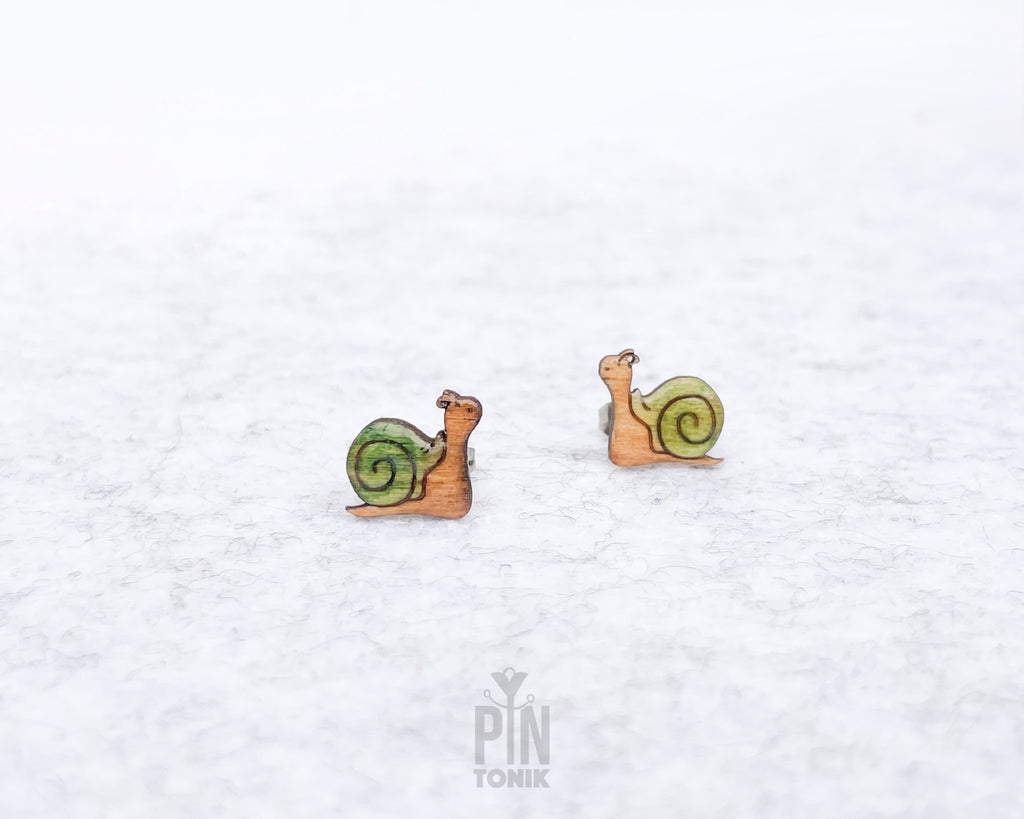 A pair of small stud earrings shaped like snails, with green and yellow spiral shells and tan bodies, photographed on a light textured white background. A small 'PIN TONIK' mark appears near the bottom center.