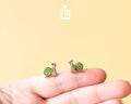A pair of small snail stud earrings resting on a person's fingertips against a soft yellow background. The earrings feature green shells with brown spiral details and pinkish-brown bodies.