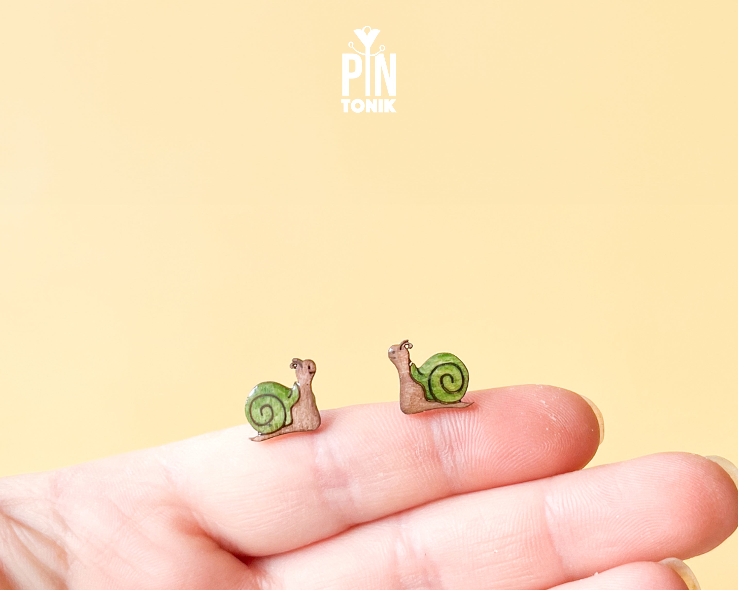 A pair of small snail stud earrings resting on a person's fingertips against a soft yellow background. The earrings feature green shells with brown spiral details and pinkish-brown bodies.