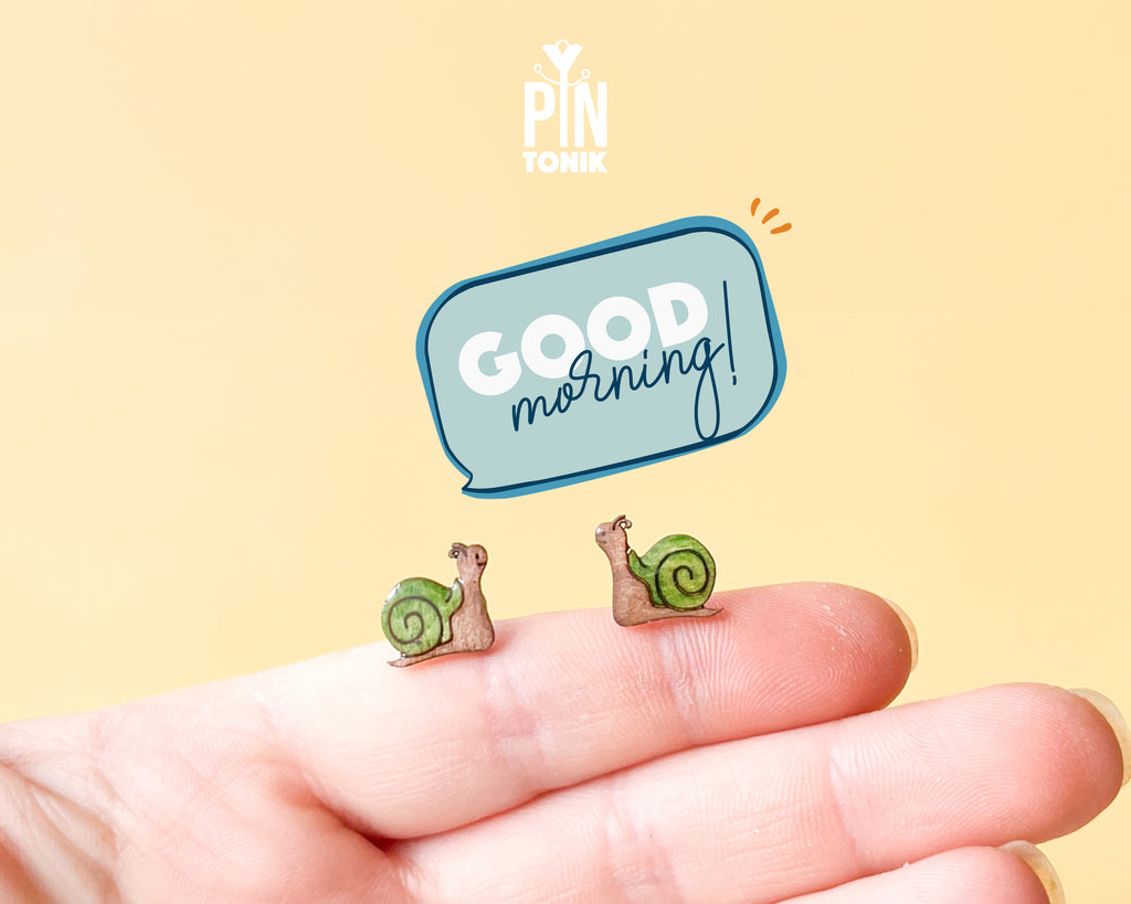 Two small illustrated snail earrings displayed on a person's fingertips against a pale yellow background, with a blue speech-bubble graphic reading 'GOOD morning!' and a 'PIN TONIK' logo above.