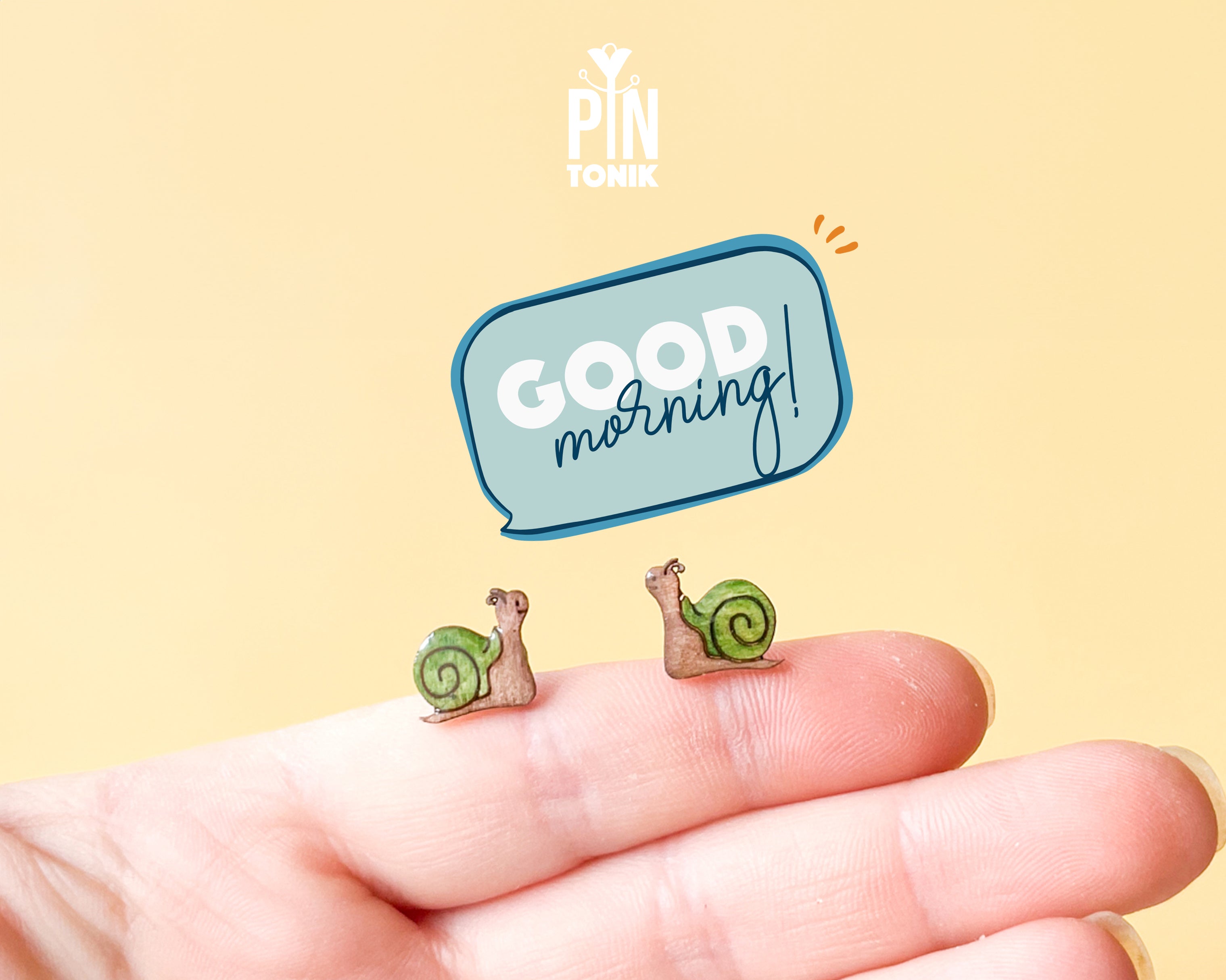 Two small illustrated snail earrings displayed on a person's fingertips against a pale yellow background, with a blue speech-bubble graphic reading 'GOOD morning!' and a 'PIN TONIK' logo above.