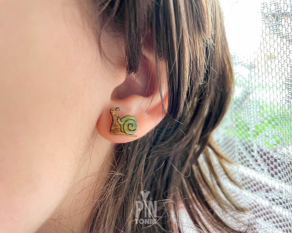 A close-up of a person's ear wearing a small snail-shaped stud earring. The earring features a brown body with a green spiral shell.
