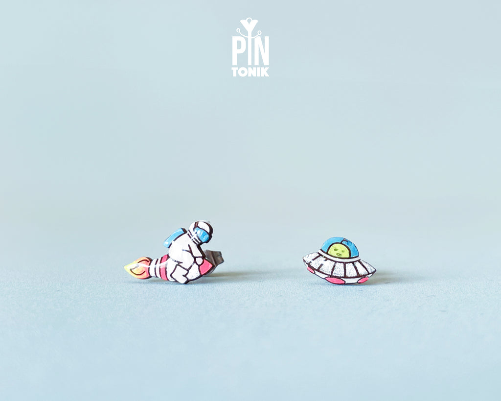 Handmade wooden mismatched stud earrings featuring an astronaut floating in space on one ear and a pastel alien UFO spaceship on the other by PinTonik.