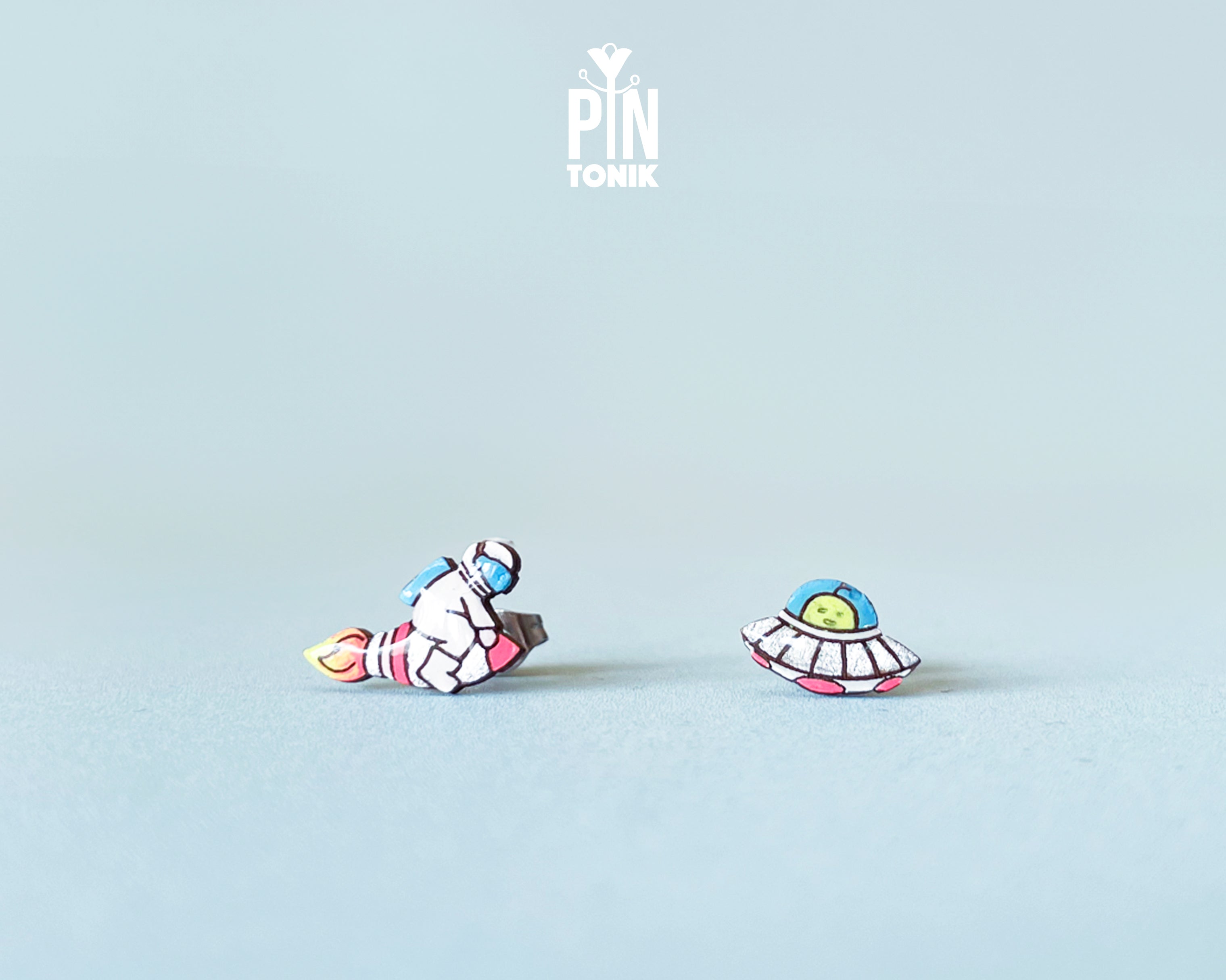 Handmade wooden mismatched stud earrings featuring an astronaut floating in space on one ear and a pastel alien UFO spaceship on the other by PinTonik.