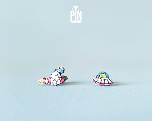 Handmade wooden mismatched stud earrings featuring an astronaut floating in space on one ear and a pastel alien UFO spaceship on the other by PinTonik.