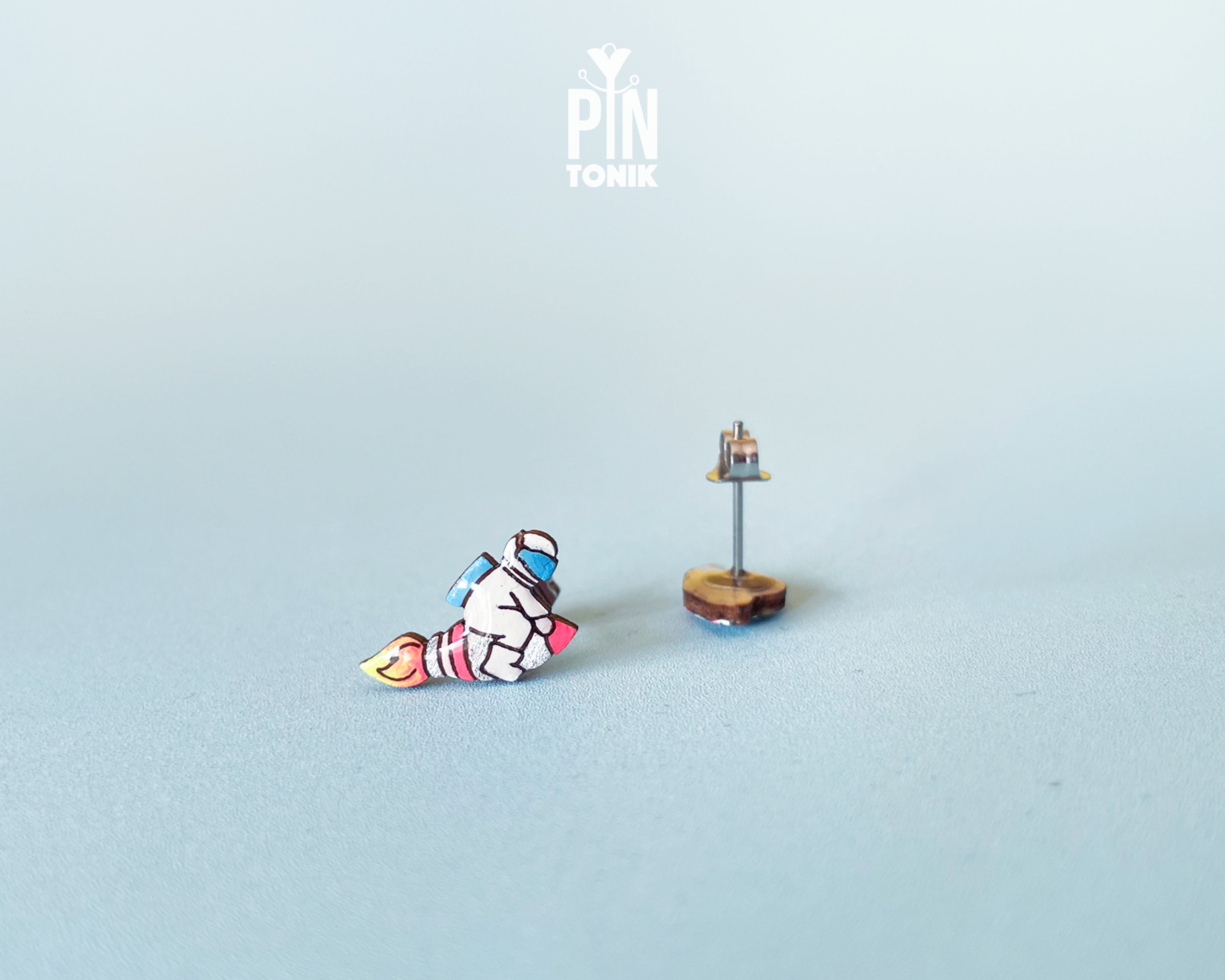 Handmade wooden mismatched stud earrings featuring an astronaut floating in space on one ear and a pastel alien UFO spaceship on the other by PinTonik.